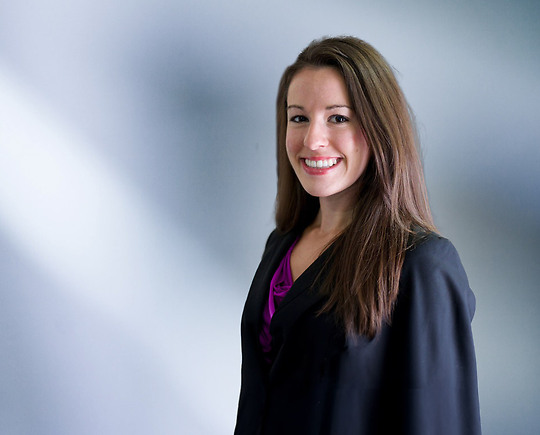 A woman, Julie Hamlet, in a black blazer and purple top is smiling at the camera, standing against a blurred blue and white background.